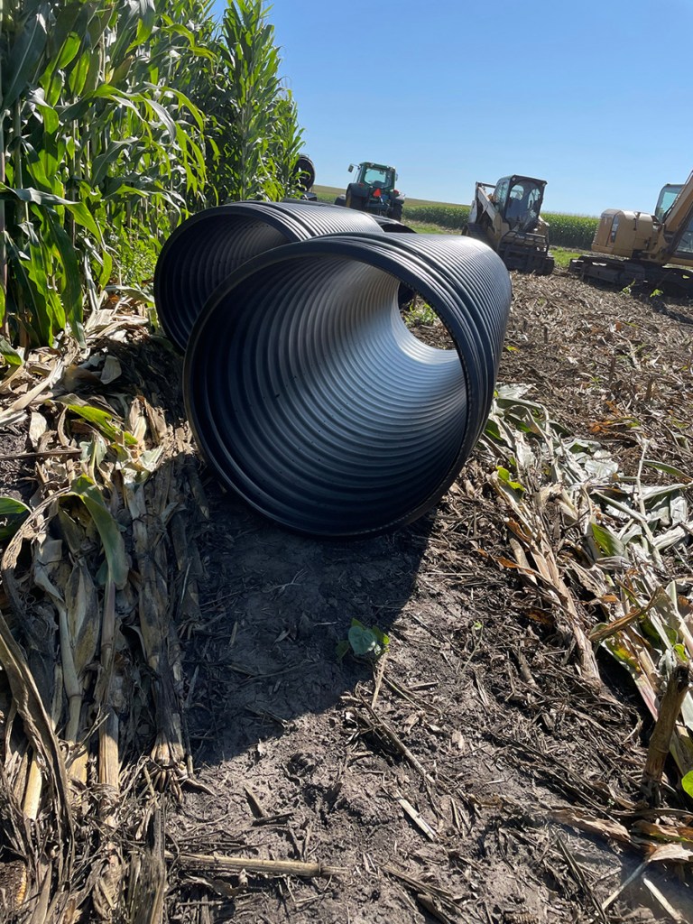 A large piece of field tile sitting in a corn field next to some heavy equipment.